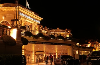 A luxurious casino at night, illuminated with warm, golden lights. The building features elegant architecture with columns and decorative elements. A few people are walking towards the entrance, and there are a couple of parked cars nearby.
