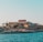 A seaside view of a large building labeled as a casino, situated on a rocky shore. The building is surrounded by palm trees and other smaller structures. The calm sea water reflects the sky, and there are several closed umbrellas lined along the waterfront.