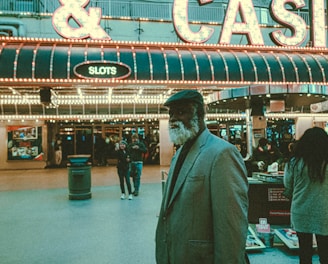 An older man with a gray beard and wearing a suit and hat stands in front of a brightly lit casino. The background features a large illuminated sign reading 'CASINO' and 'SLOTS' among other words, with numerous lights outlining the entrance. Several other people are visible in the background, engaged in various activities, some walking, some standing, and one looking at something on display.
