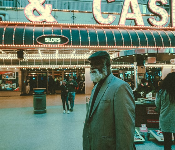 An older man with a gray beard and wearing a suit and hat stands in front of a brightly lit casino. The background features a large illuminated sign reading 'CASINO' and 'SLOTS' among other words, with numerous lights outlining the entrance. Several other people are visible in the background, engaged in various activities, some walking, some standing, and one looking at something on display.